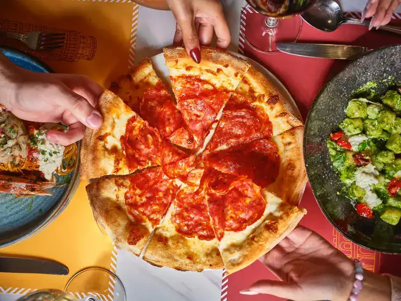 Hands reaching for slices of a pepperoni pizza in the middle of the table