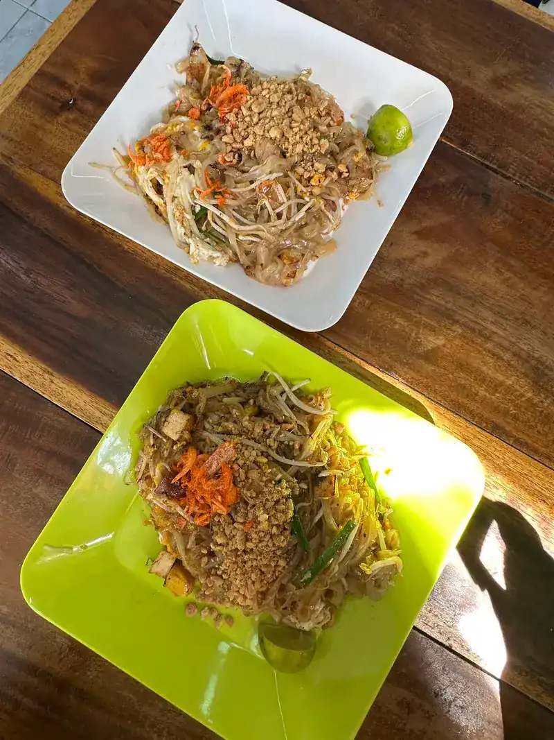 Two plates of pad thai on a wooden table, one on a white plate and one on a green plate