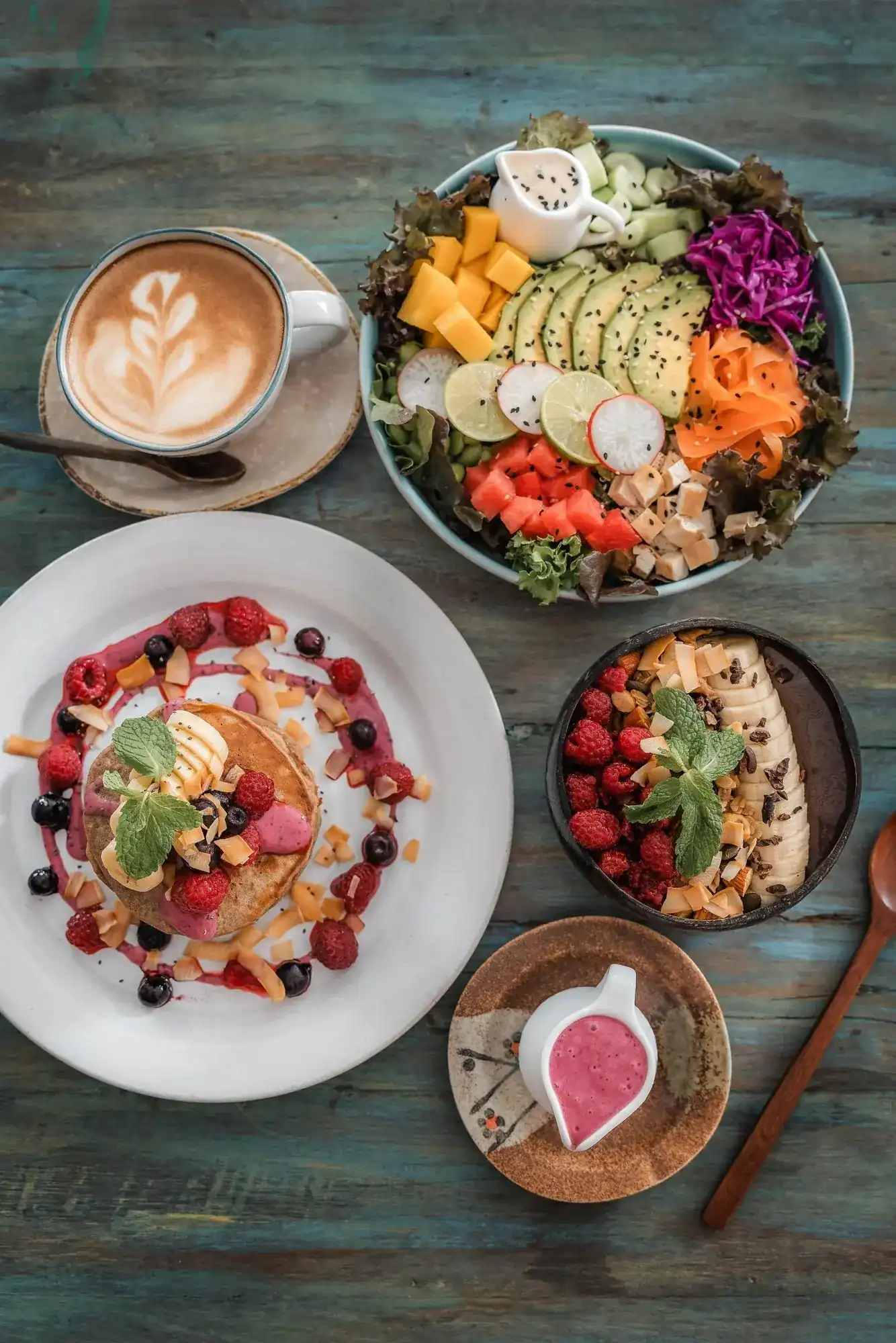 A weathered turquoise table seen from above: pancakes with berries, a salad bowl, a fruit bowl and a coffee