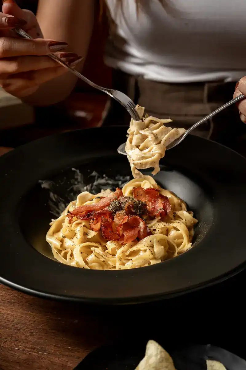 Fettuccine in cream sauce with crisp bacon, lifted with a fork and spoon from a black bowl