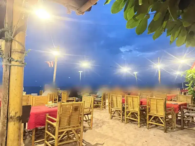Bamboo chairs and red tables on the sand under bright lights after dark, the sea behind