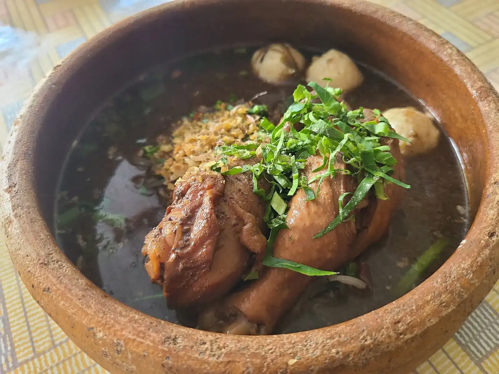 A clay pot of boat noodle soup with a chicken drumstick, meatballs and fresh greens on top