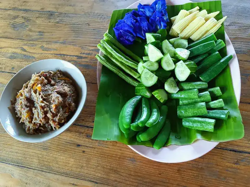 Fresh greens on a banana leaf with butterfly pea flowers, baby corn, okra and cucumber, beside a bowl of chilli relish
