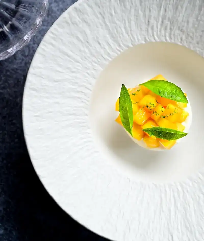 A white textured plate with diced yellow fruit and green leaves, seen from above