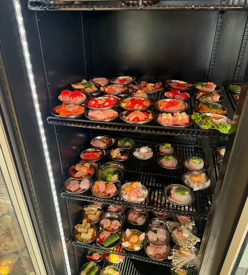 Small steel plates of dim sum lined up on the shelves of a glass cabinet, ready for the steamer