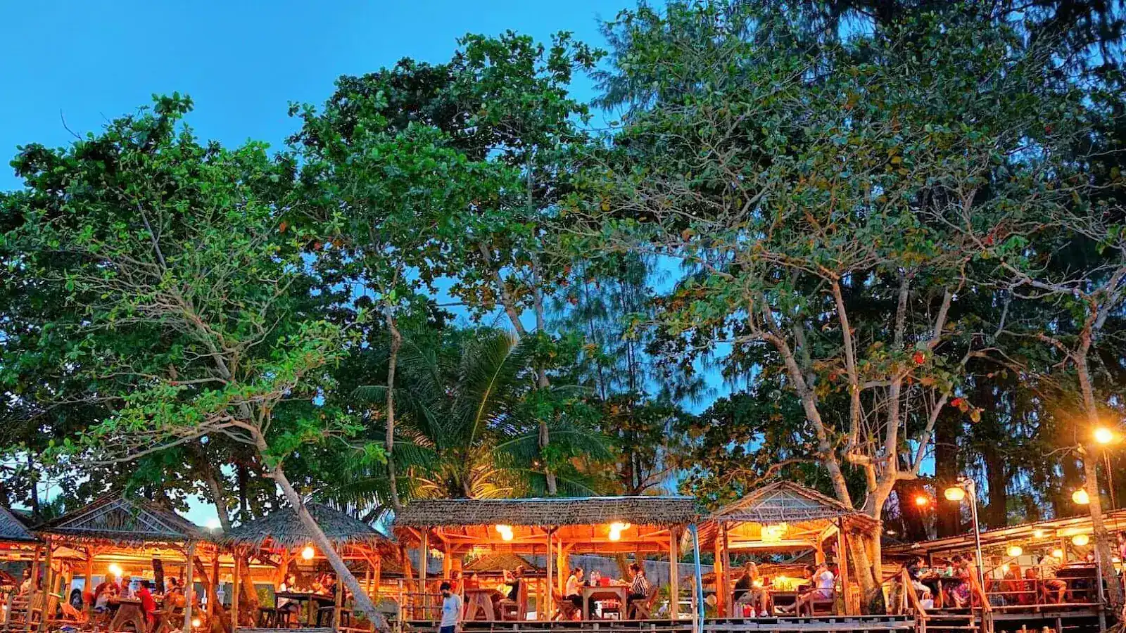 Thatched salas lit up under tall trees at dusk, with guests at the tables