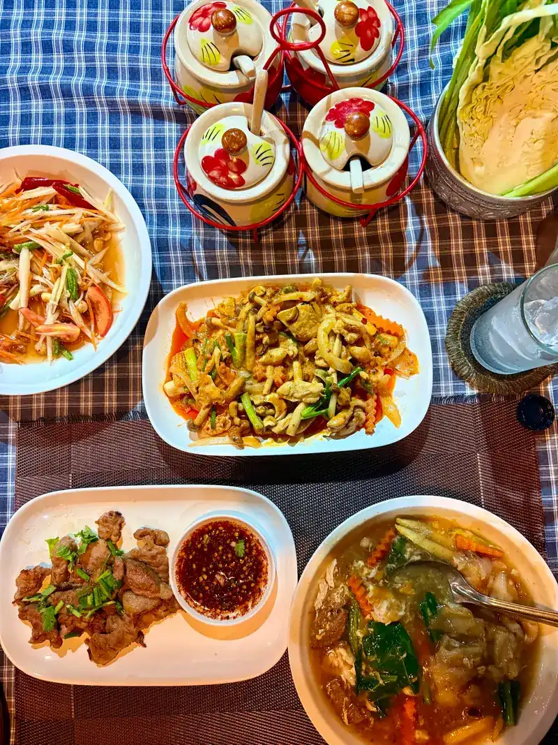 Seen from above: papaya salad, a stir-fry, sliced grilled meat with dipping sauce and a bowl of vegetables in gravy, beside painted condiment jars and a bowl of cabbage and greens