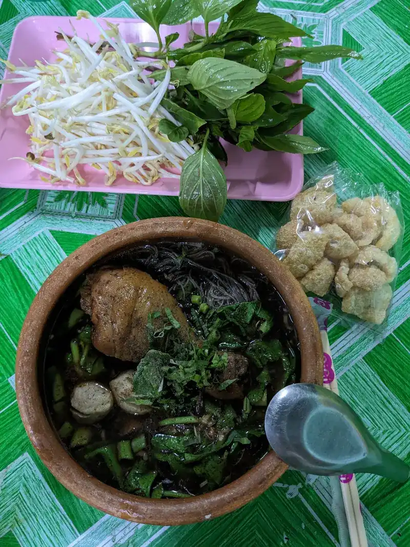 Boat noodles in a clay pot with meatballs and chicken, next to a plate of bean sprouts and fresh herbs and a bag of pork crackling