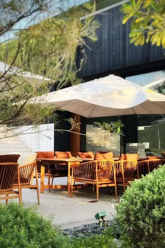 Garden terrace with timber chairs, rust-coloured cushions and a white parasol in front of the dark timber building