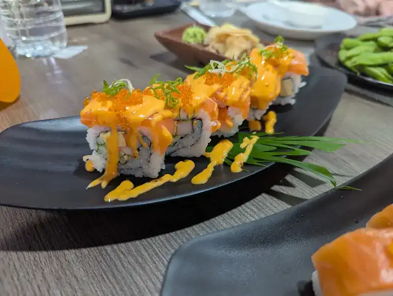 A maki roll topped with salmon, orange sauce, fish roe and spring onion on a black plate