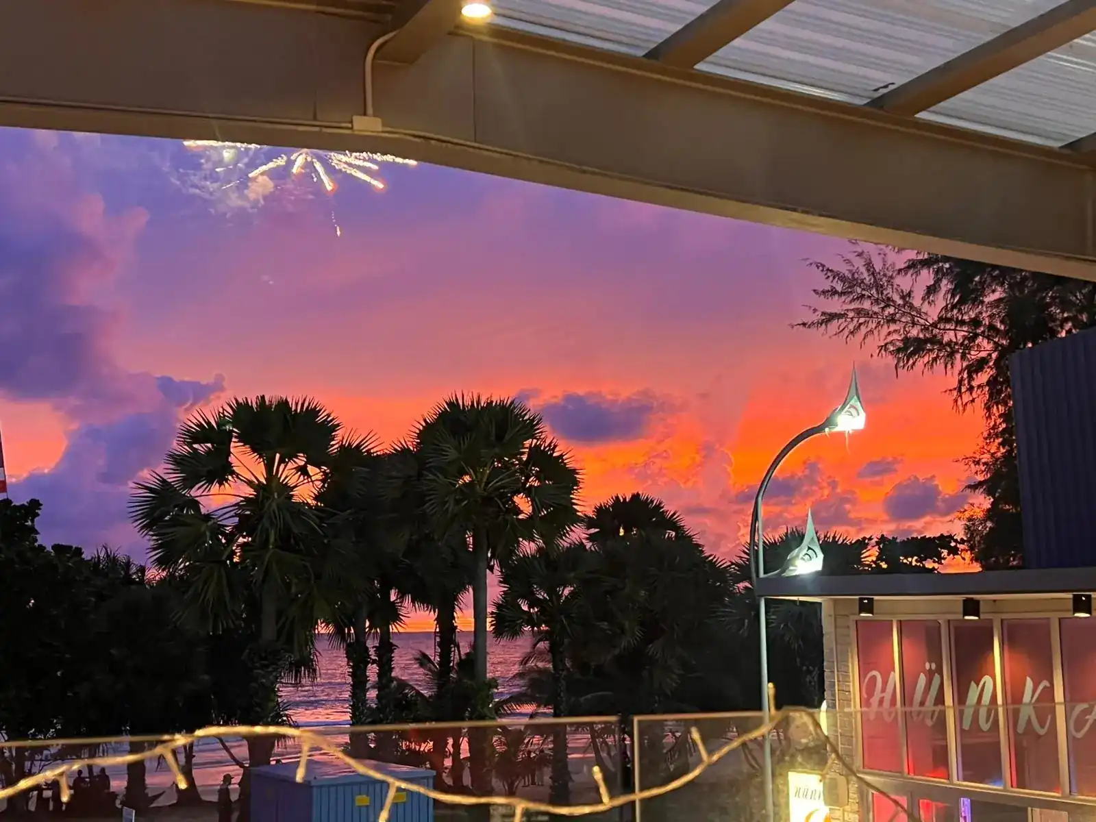 Sunset over Karon Beach: palm trees and the sea across the road from Hünkâr, under a pink and orange sky