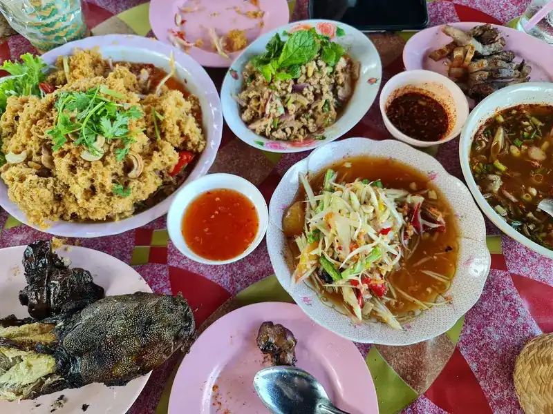 A full table of Isaan dishes: papaya salad, larb, a spicy soup, grilled fish, grilled meat and chilli dips, with a sticky rice basket