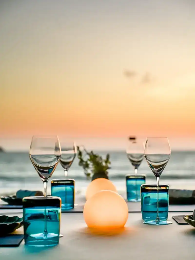 Wine glasses, blue tumblers and glowing lamps on a table by the sea at dusk