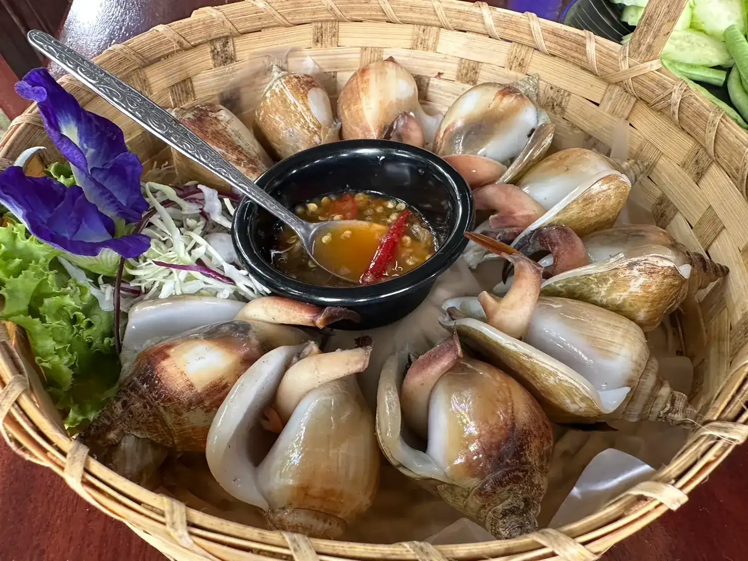 Dog conch served in a woven basket with seafood dipping sauce and a butterfly pea flower