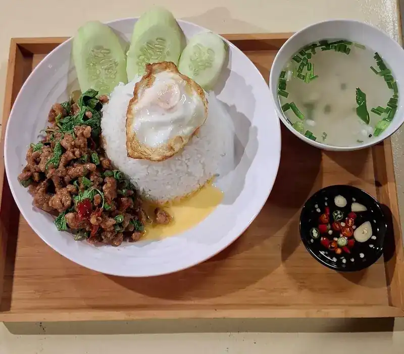 Holy basil stir-fry on rice with a fried egg and cucumber, a cup of clear soup and chilli fish sauce, on a wooden tray
