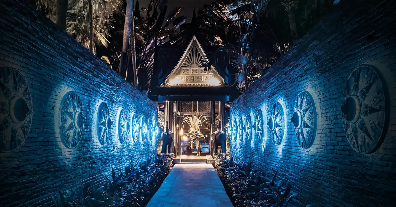 A path between brick walls lined with blue-lit carved shields, leading to the gabled wooden house of Black Ginger at night