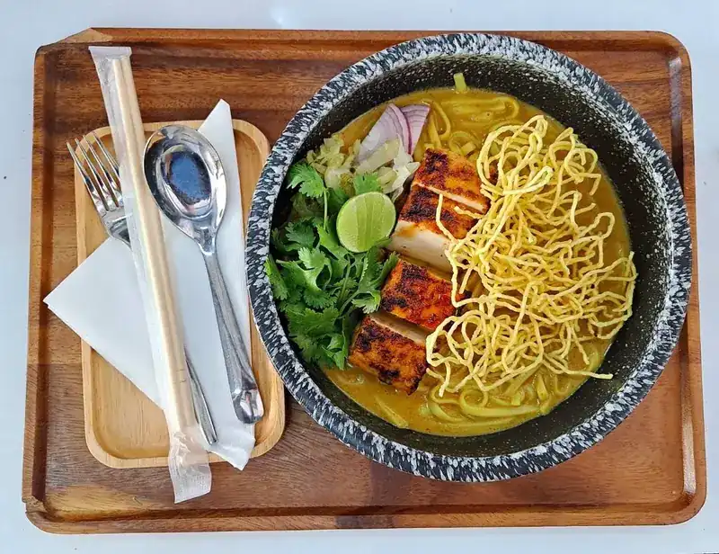 Khao soi topped with crispy noodles, with lime and coriander on a wooden tray
