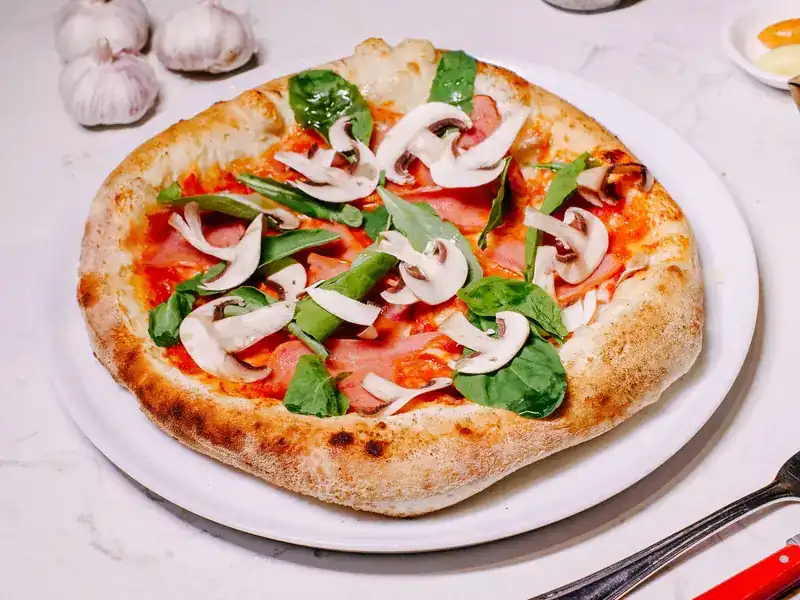 A pizza with ham, sliced mushrooms and spinach leaves on a marble table