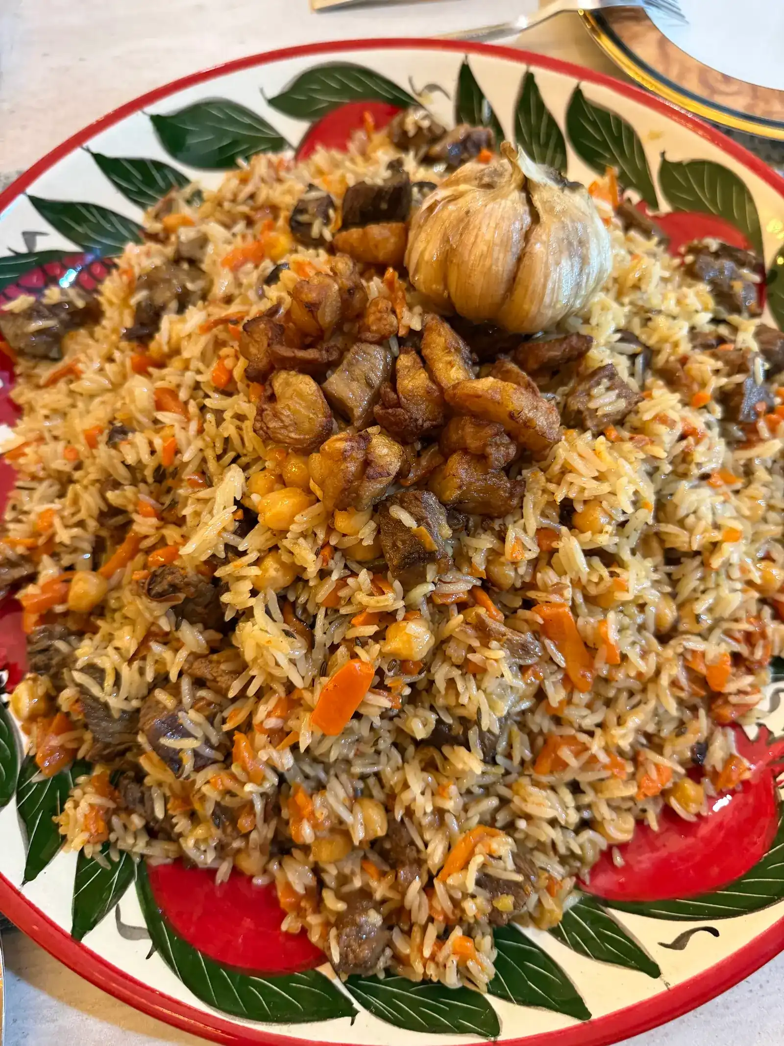 Plov piled on a red and green painted plate: rice, carrot, chickpeas and meat with a whole head of garlic