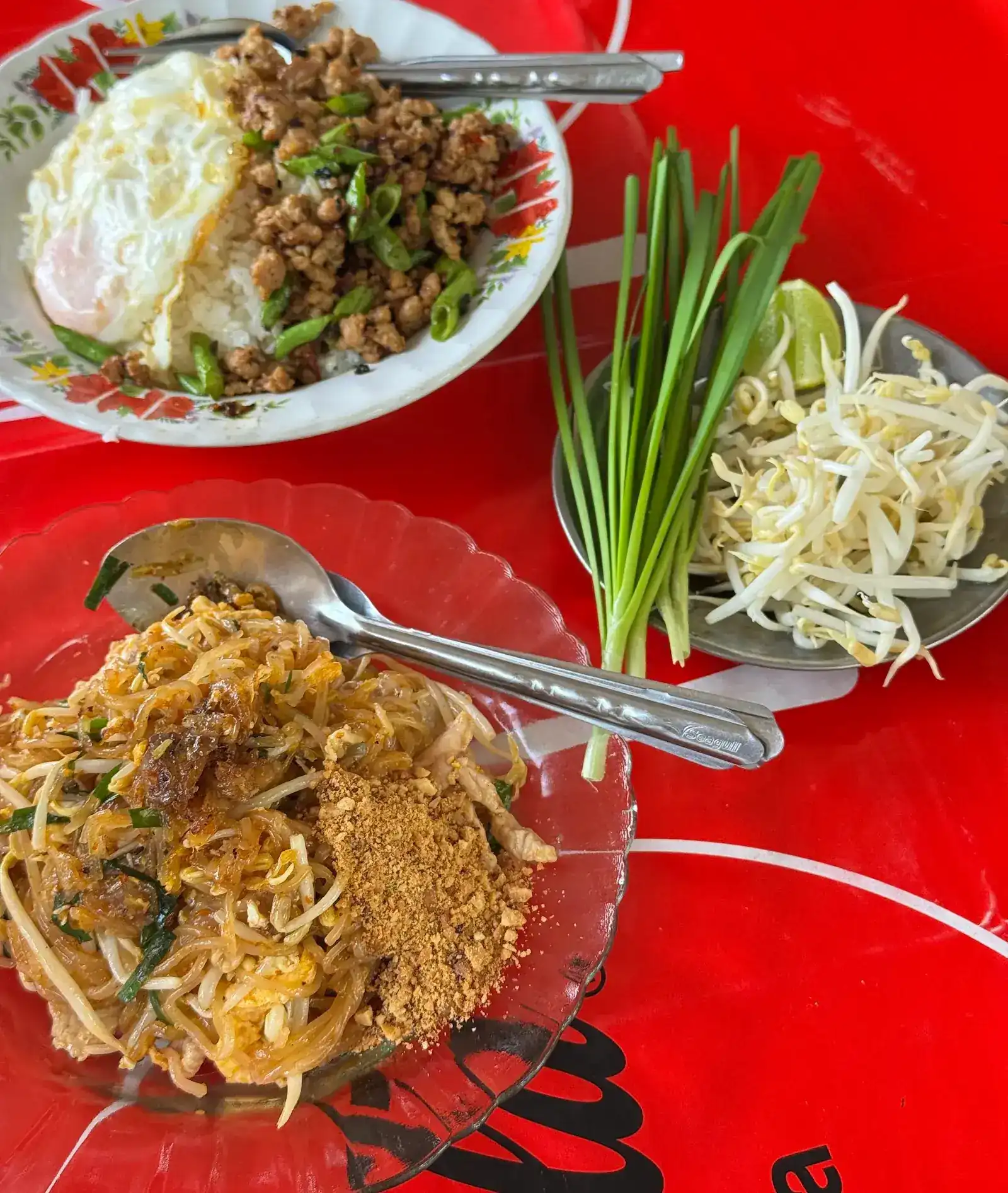 Pad thai with ground peanuts on a glass plate, minced pork with long beans over rice with a fried egg, and a side plate of bean sprouts, Chinese chives and lime on a red tablecloth