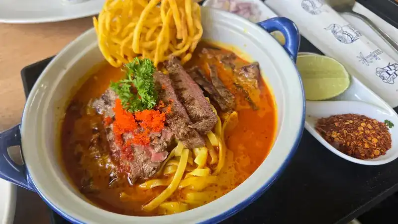 Curry noodle soup with slices of grilled beef, crispy noodles and parsley on top, with lime and chilli flakes on the side