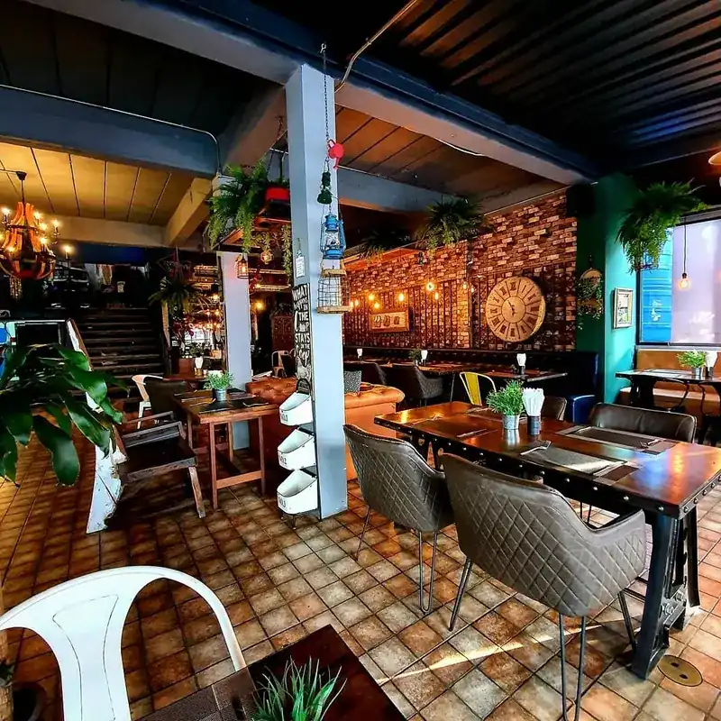 The dining room: tiled floor, ferns hanging from the beams, a brick wall, a teal wall and a large wall clock. ห้องอาหาร พื้นปูกระเบื้อง ต้นเฟิร์นห้อยจากคาน ผนังอิฐ ผนังสีเขียวหัวเป็ด และนาฬิกาแขวนเรือนใหญ่