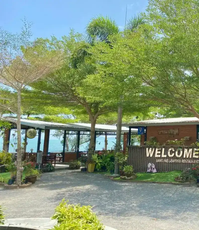 The entrance and its welcome sign under shade trees, the sea visible behind
