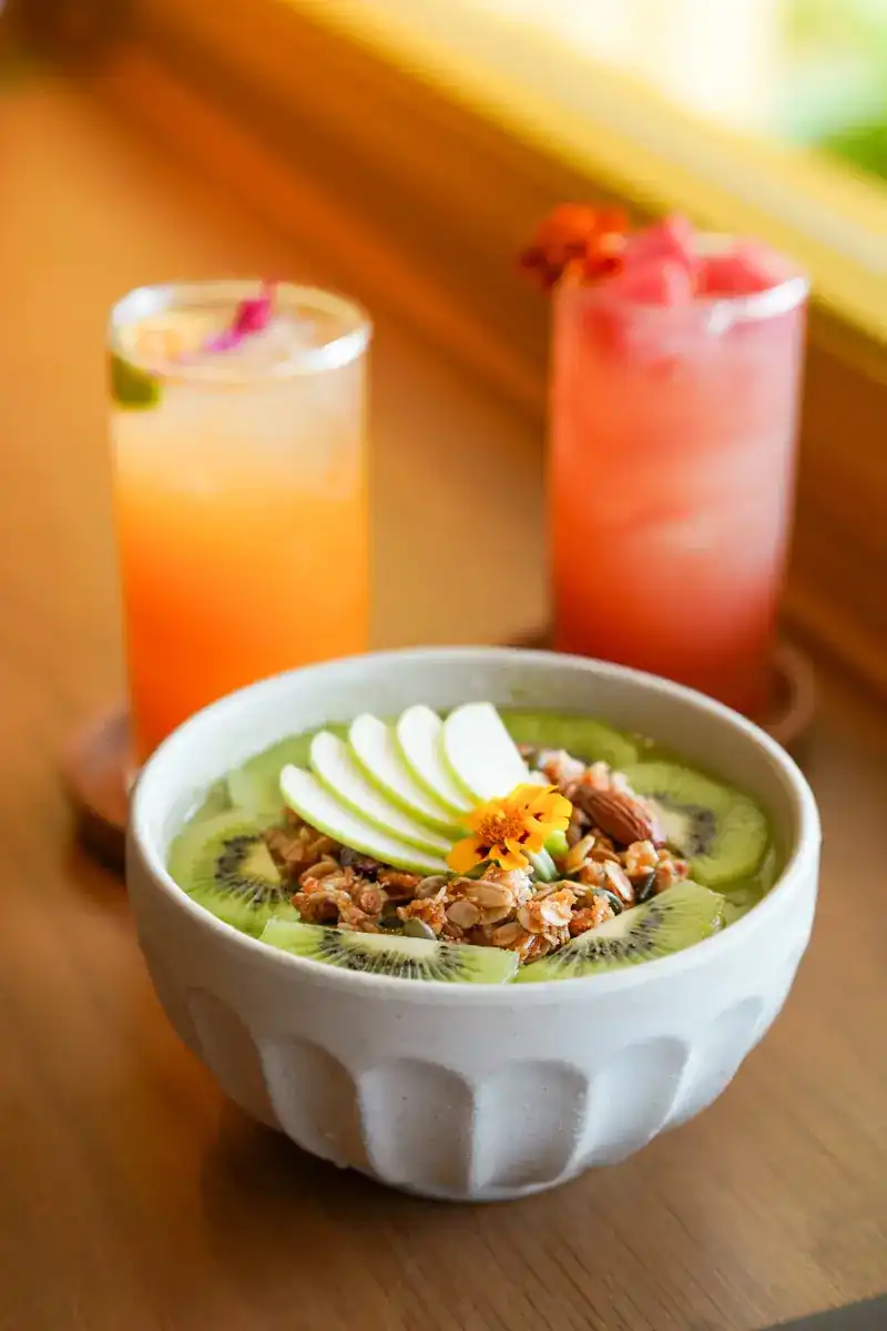 A bowl of granola with kiwi and apple slices, two fruit drinks behind