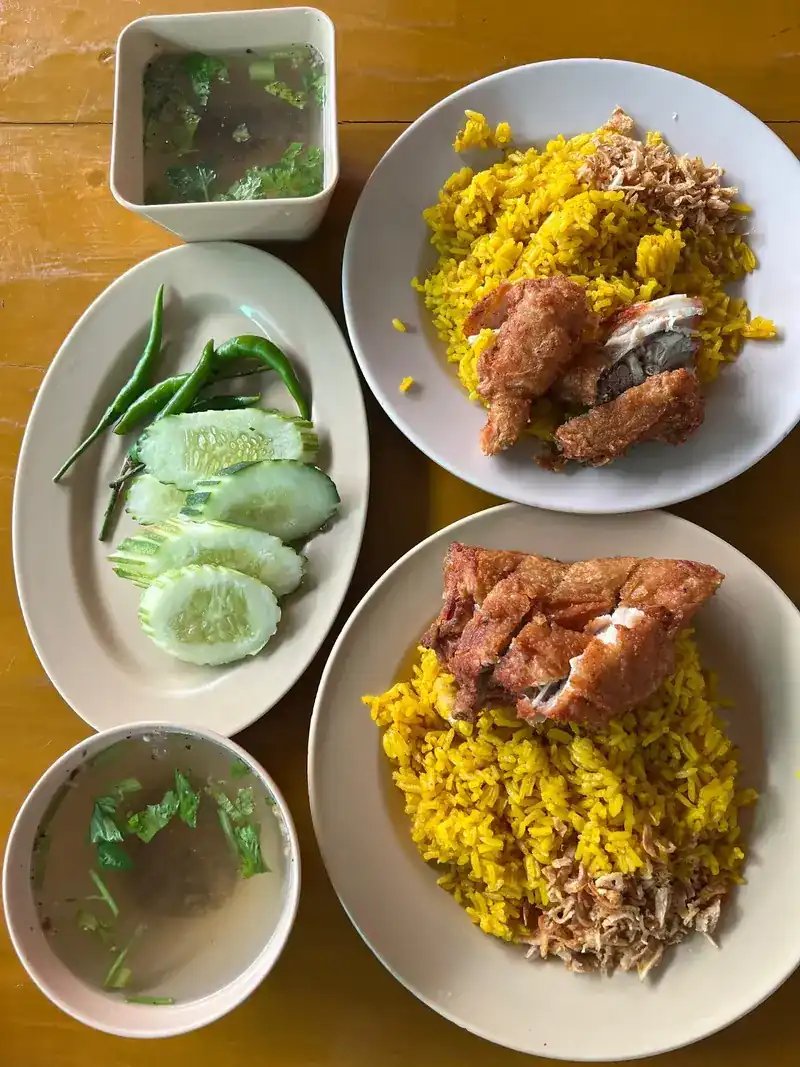 Two plates of yellow rice with fried chicken, two bowls of clear soup and a plate of cucumber and chilli, seen from above