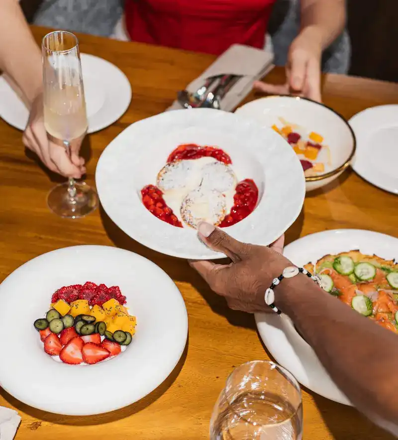 A plate of syrniki with berry sauce passed across a table set with fruit and salmon