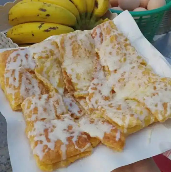 Roti cut into squares under sweetened condensed milk, with bananas and eggs behind