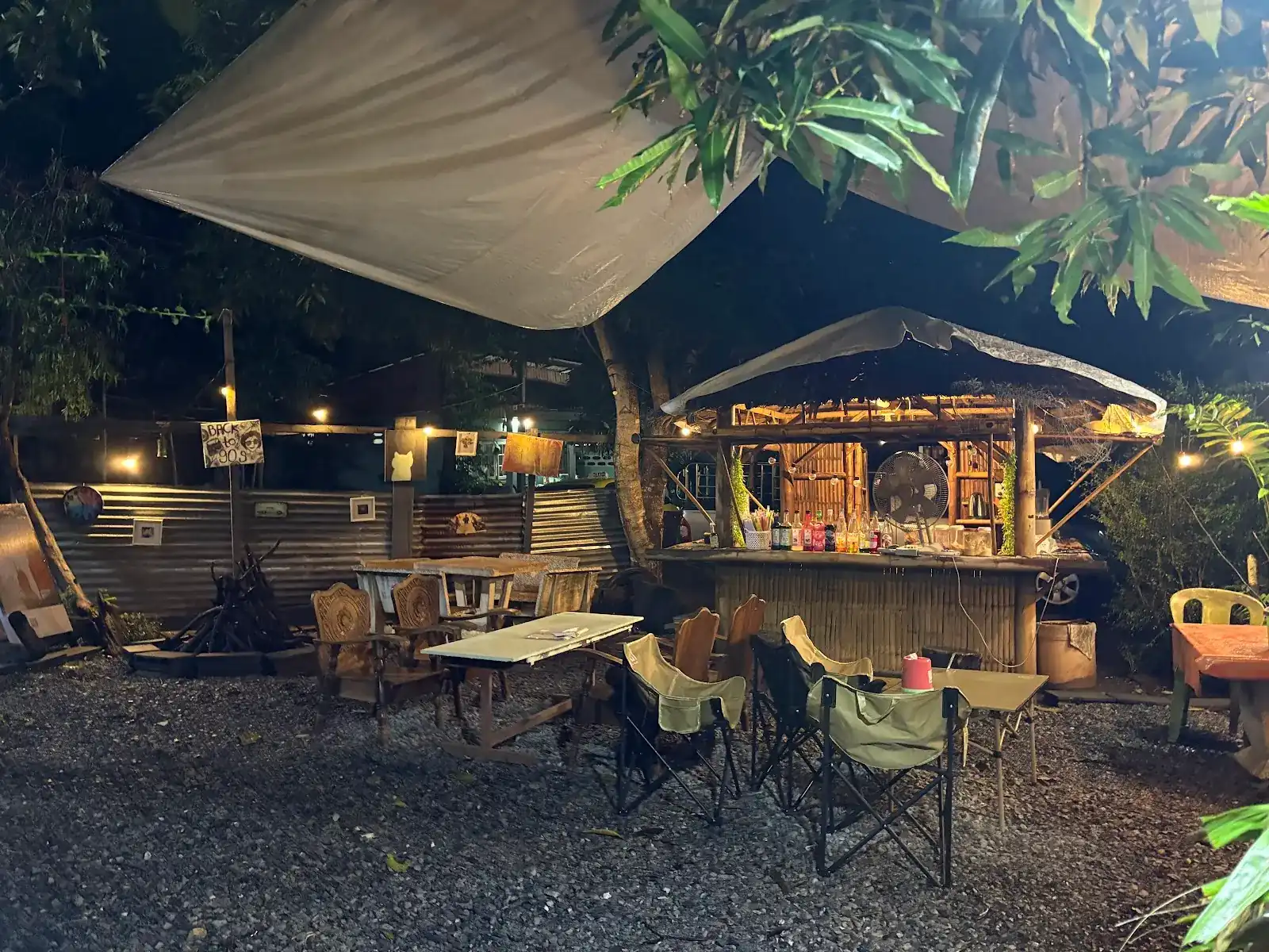 The garden at night: a canvas sail over gravel, camp chairs, carved wooden chairs and a lit bamboo bar under the trees