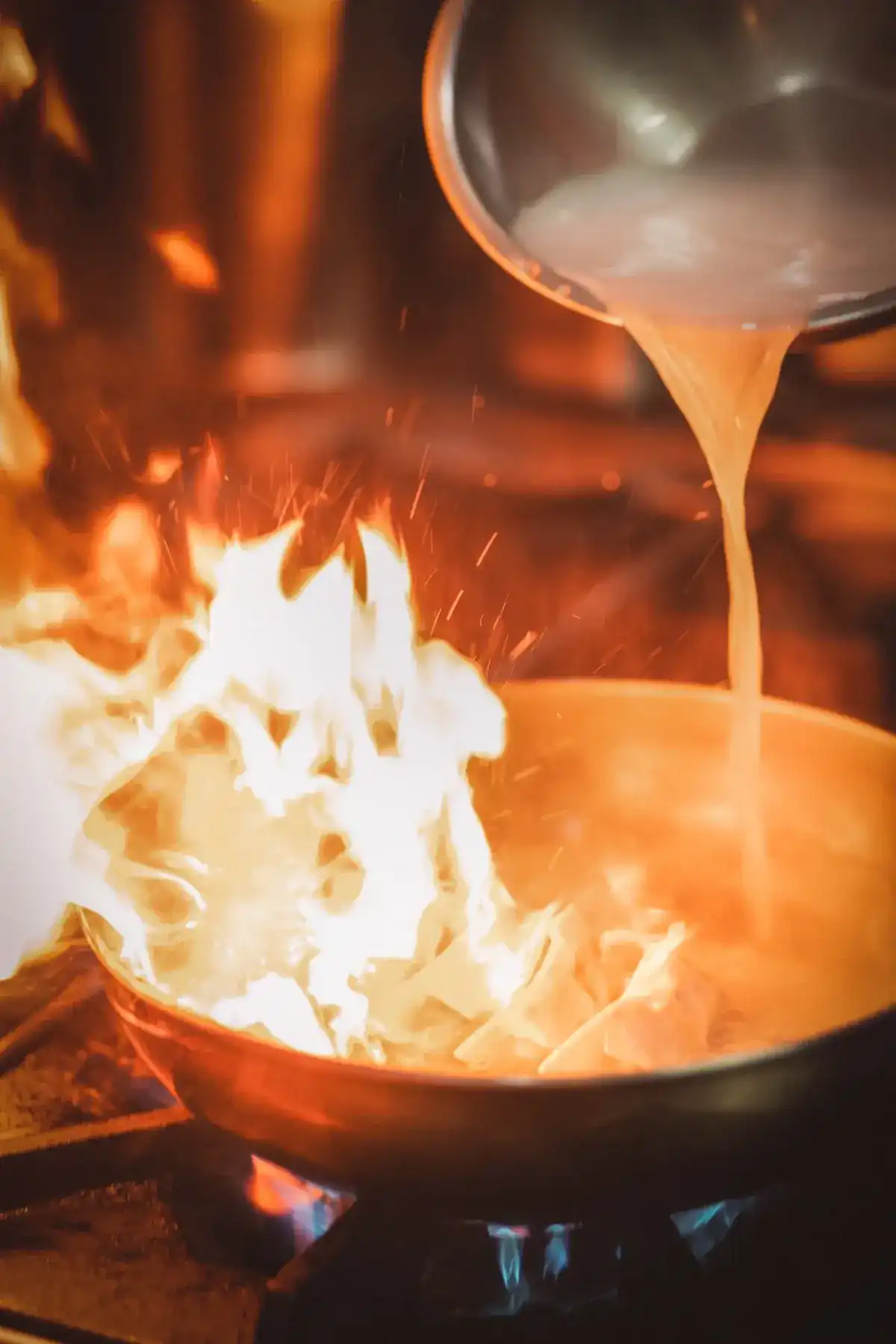Flames leap from a wok as stock is poured in from a ladle