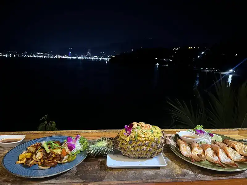 Dinner on the railing at night: a stir-fry, pineapple rice and large prawns, with the lights of the bay behind
