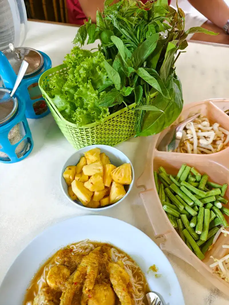 Rice noodles with curry beside a basket of fresh herbs, long beans and pineapple