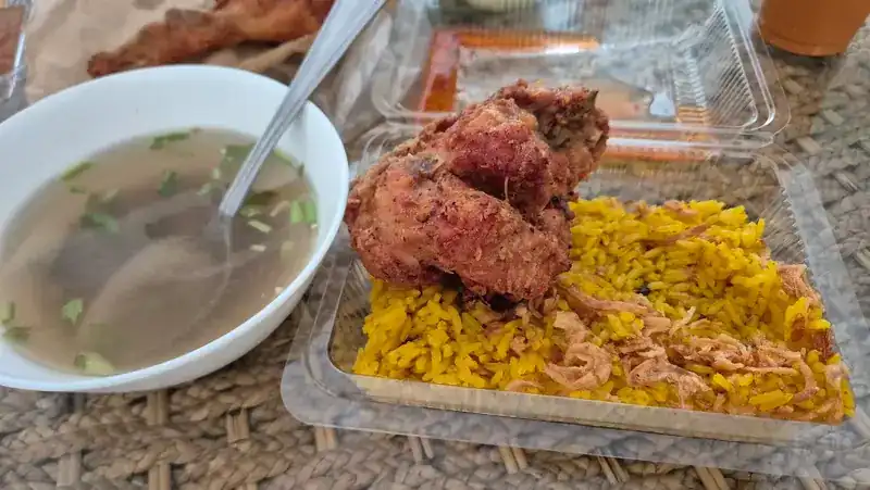 A takeaway box of yellow rice with fried chicken, next to a bowl of clear soup