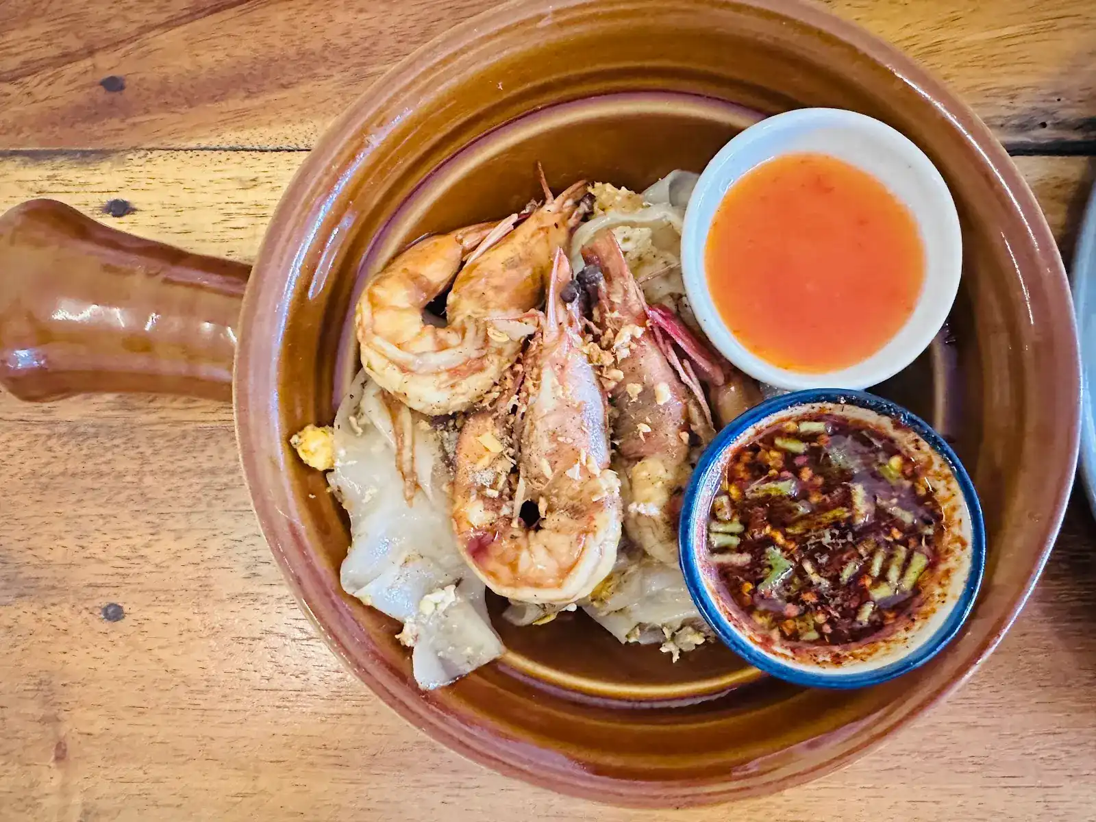 Prawns on wide rice noodles in a brown clay pan, with two small bowls of dipping sauce