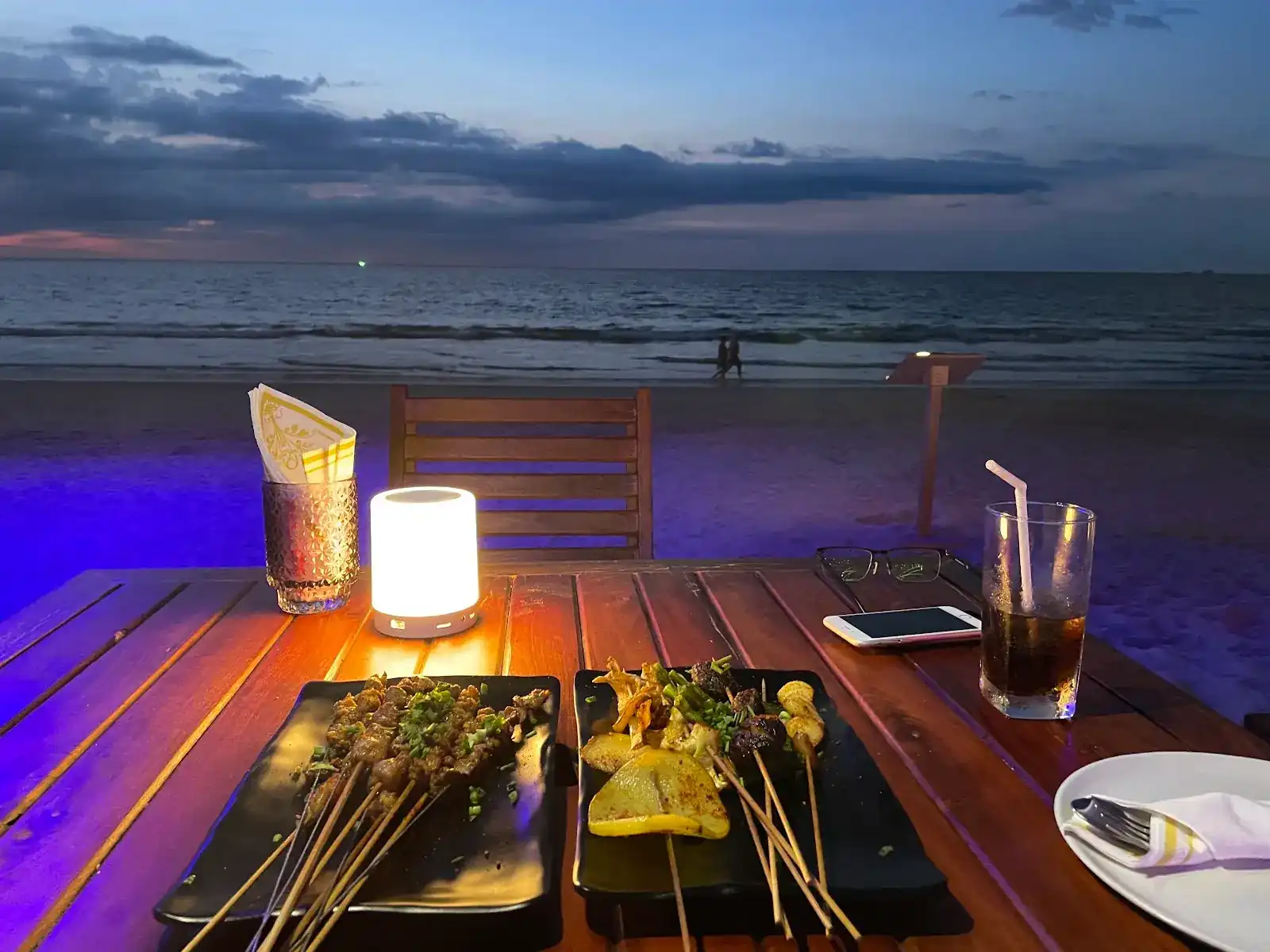 Grilled skewers and a glowing lamp on a wooden table on the sand at dusk, Bang Tao Beach