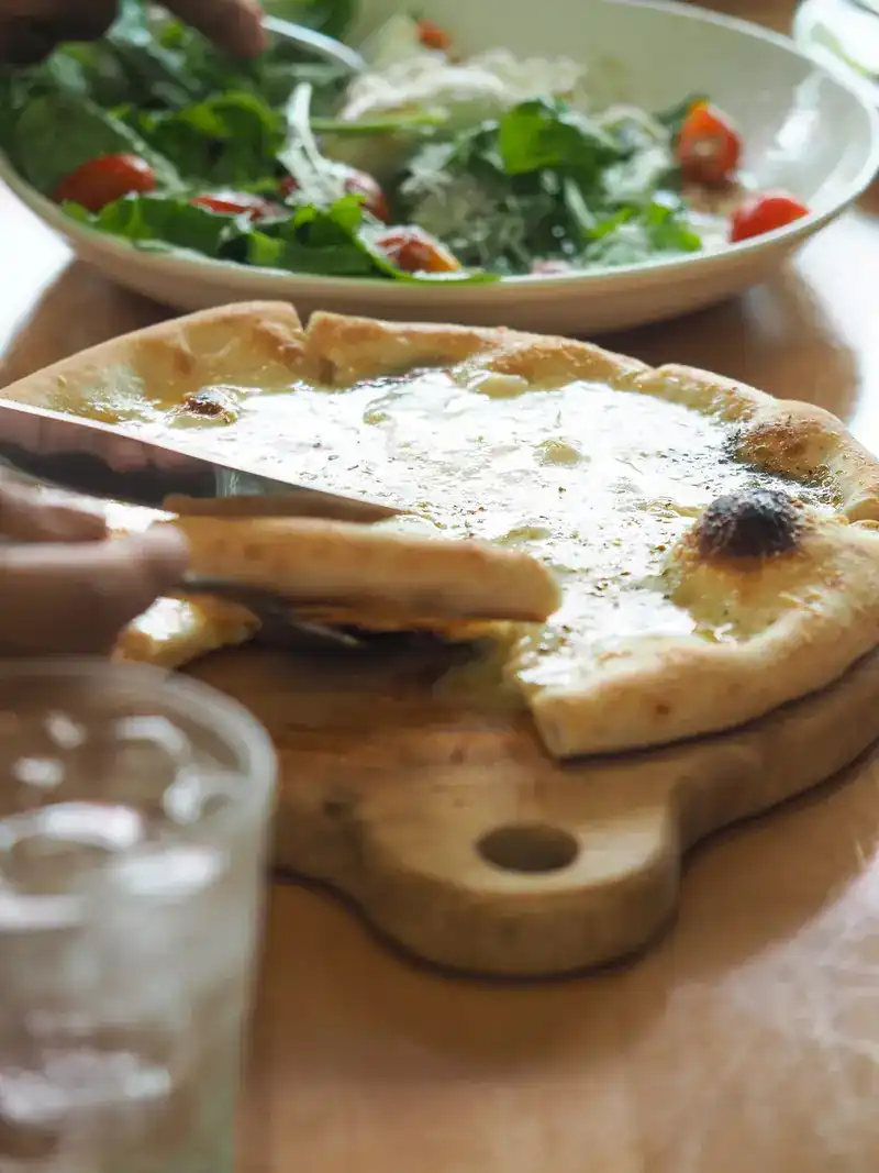 A knife cutting a white pizza on a wooden board, a green salad behind
