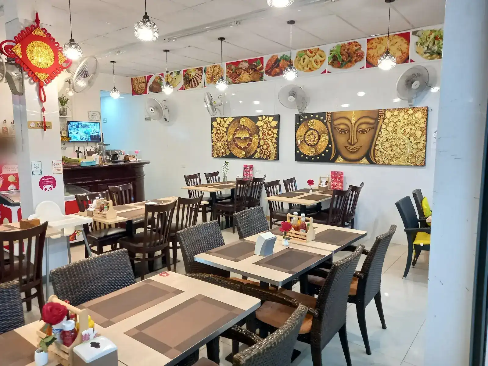 Inside the restaurant: tables with rattan and wooden chairs, pendant lamps and golden Thai paintings on the white wall / ภายในร้าน โต๊ะกับเก้าอี้หวายและเก้าอี้ไม้ โคมไฟห้อย และภาพวาดไทยสีทองบนผนังสีขาว