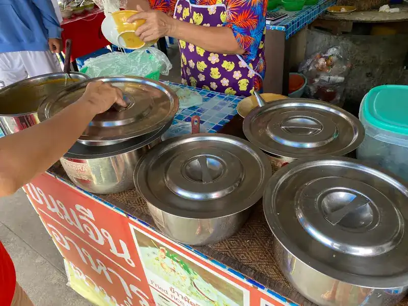 Steel curry pots with lids on the counter while a plate of noodles is served