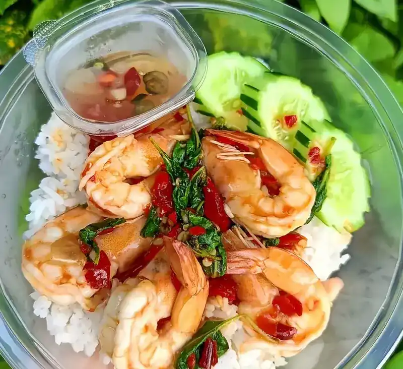 Shrimp kaprao over rice in a takeaway box, with cucumber and a small cup of chilli sauce