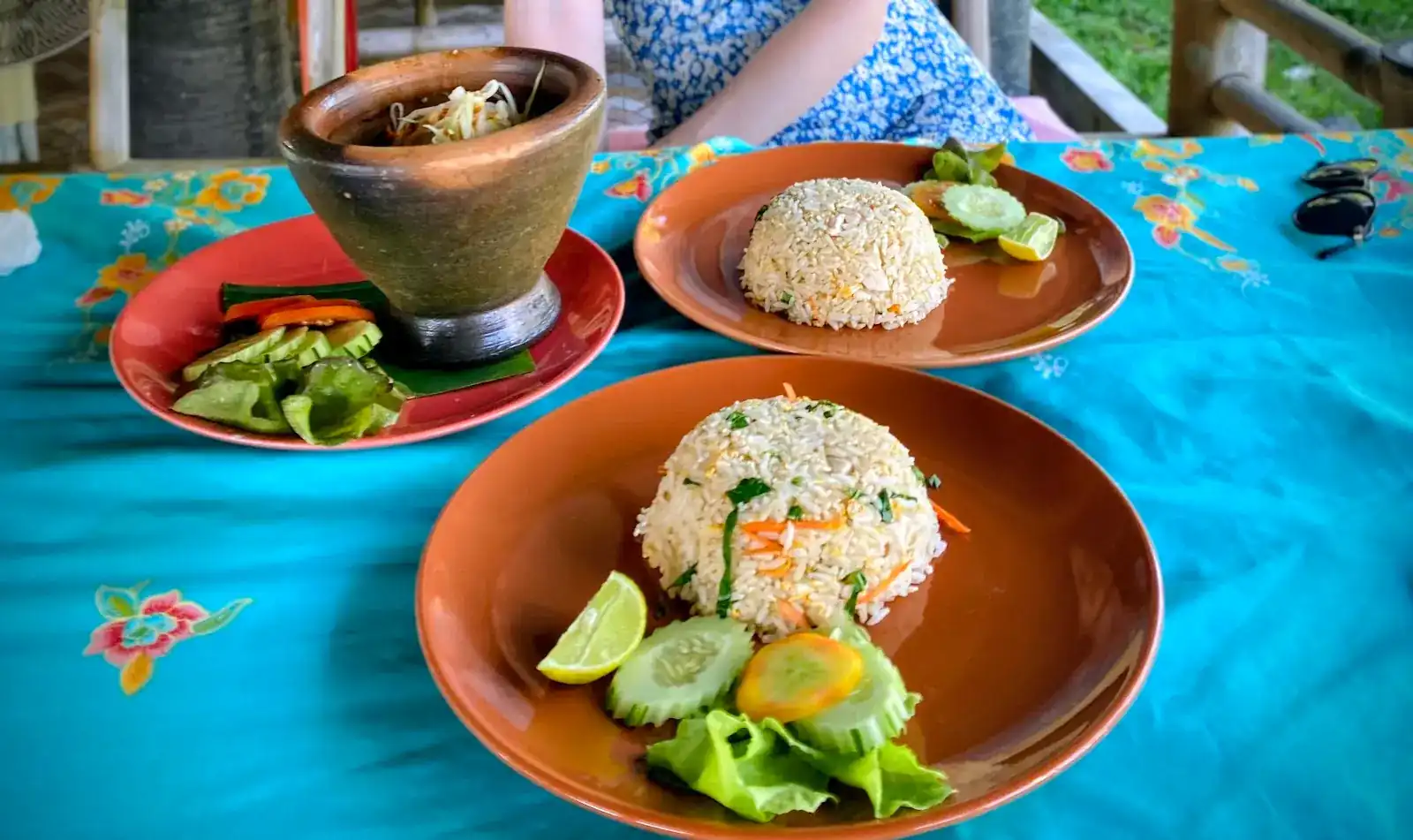 Two plates of fried rice and papaya salad in a clay mortar, on a turquoise flowered tablecloth