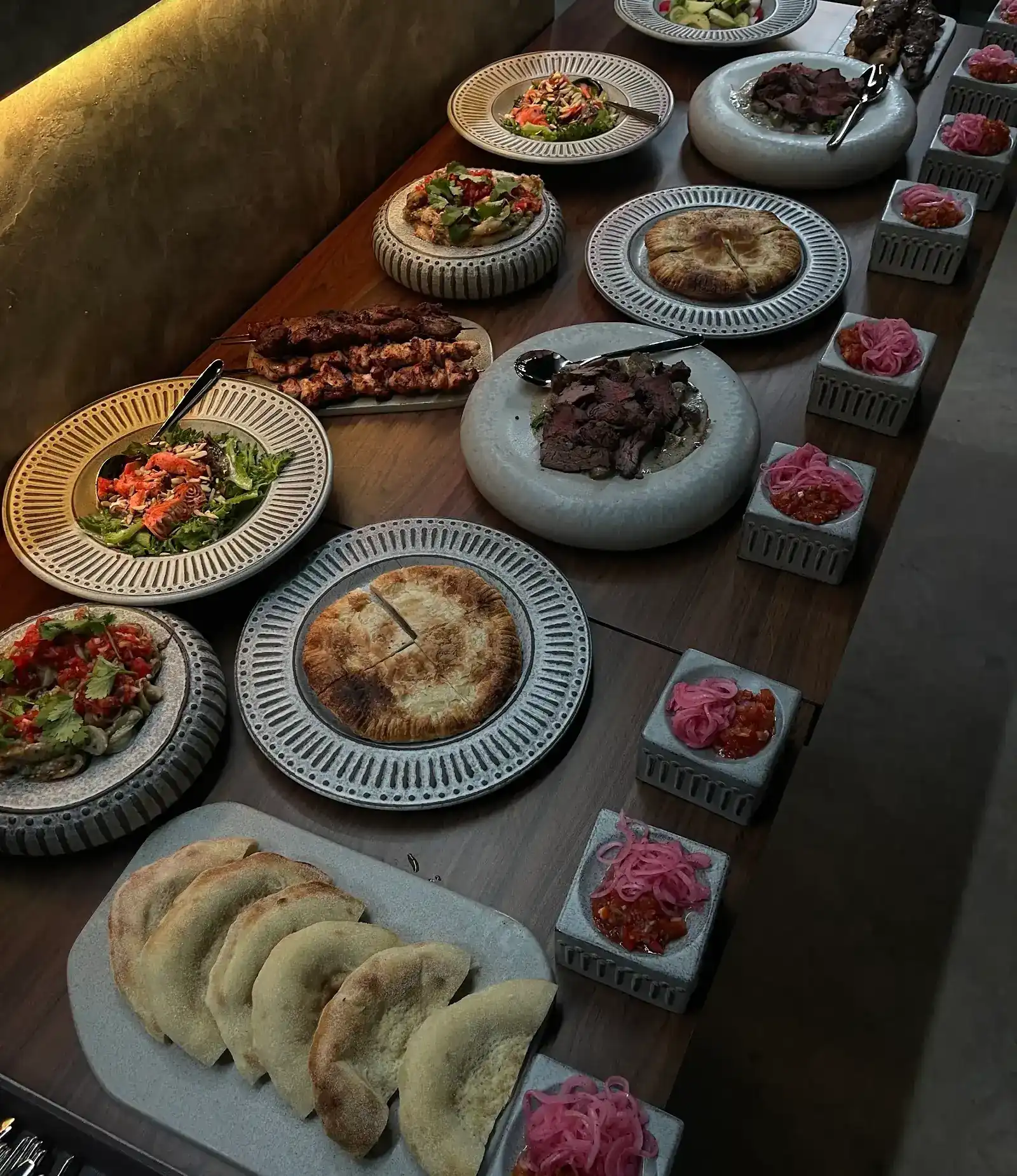 A long table set with shashlik skewers, salads, sliced roast meat, golden pies, flatbreads and small bowls of pickled onion