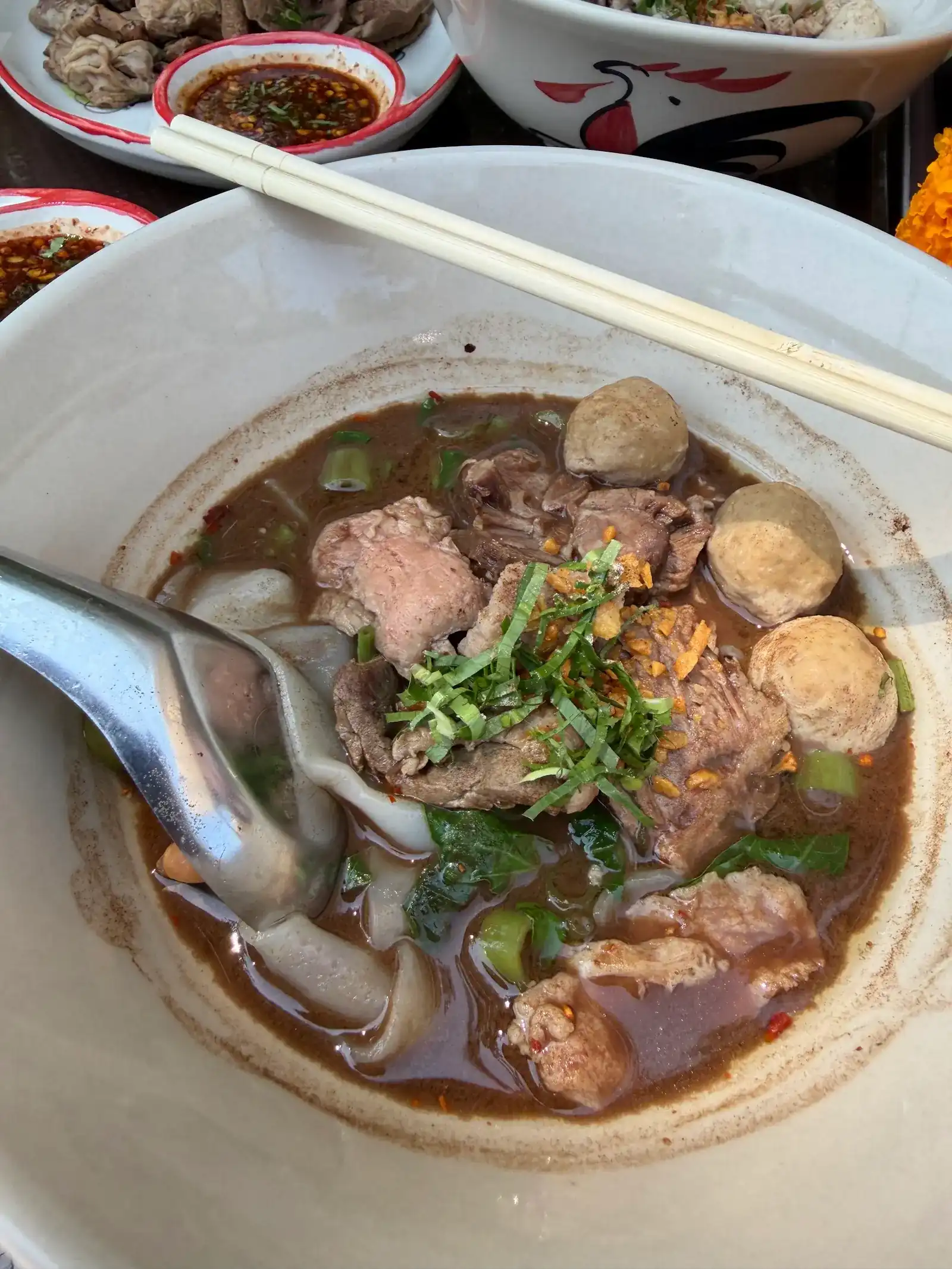 A bowl of boat noodles in dark broth with sliced meat, meatballs, greens and fried garlic, chopsticks across the rim