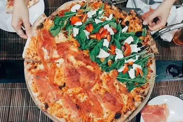 Hands reaching for a pizza that is half prosciutto crudo, half caprese with rocket