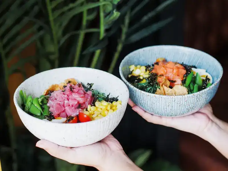 Two bowls held up in front of palm leaves, one with tuna and one with salmon, with sweetcorn, greens and seaweed