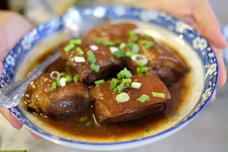 Hands holding a blue and white bowl of braised pork belly with spring onion