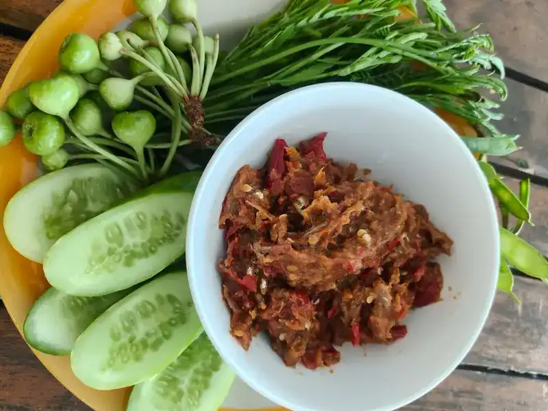 A bowl of chilli paste with cucumber, small green aubergines and fresh greens