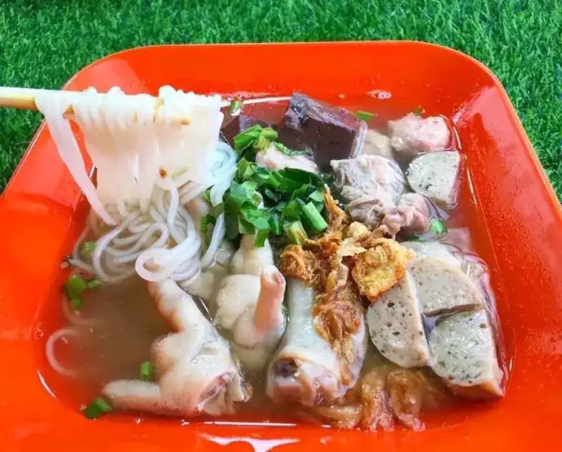 Noodles lifted with chopsticks from an orange bowl of kuay jap with chicken feet, pork balls and Vietnamese pork sausage